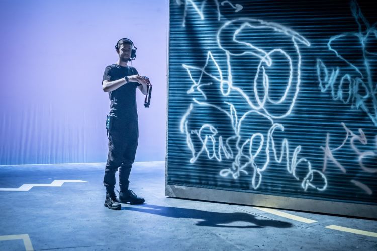 Stage Management student with a headset on is preparing the stage for the performance of Dennis of Penge. The student is standing next to a wall of graffiti which was made by Design Realisation Students.